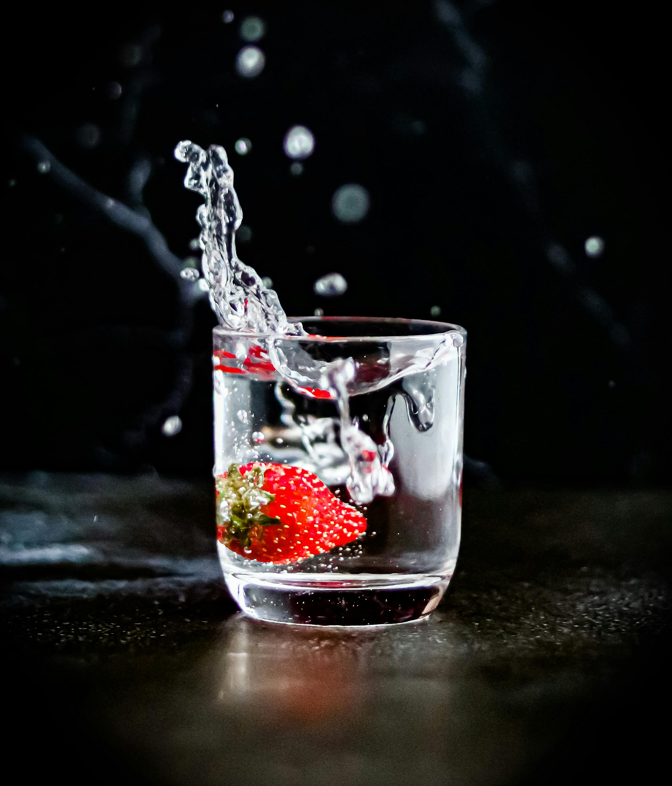 Dynamic image of a fresh strawberry splashing into a clear glass of water with droplets against a black background.