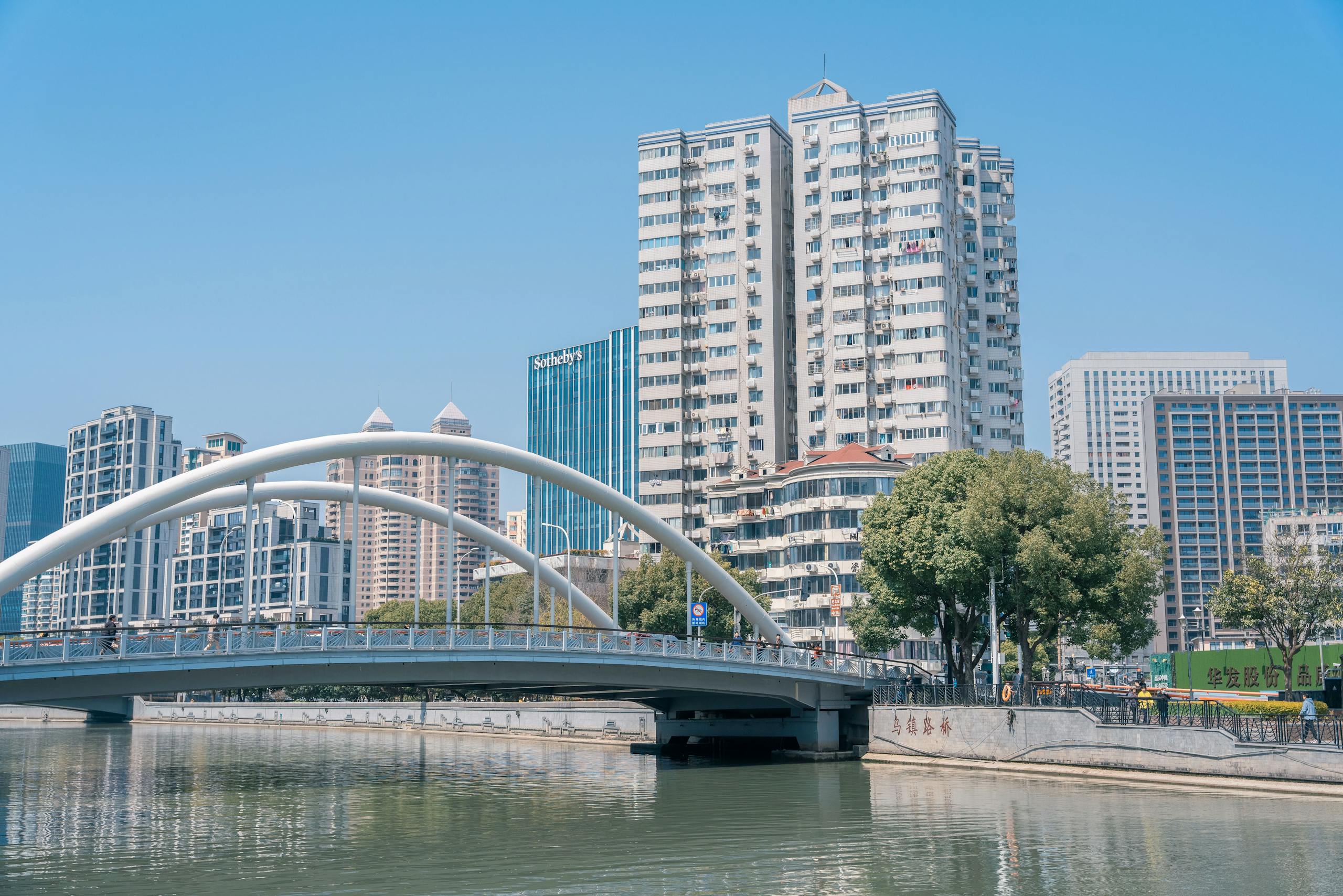 Modern cityscape with a white arch bridge crossing a river against a skyline backdrop.