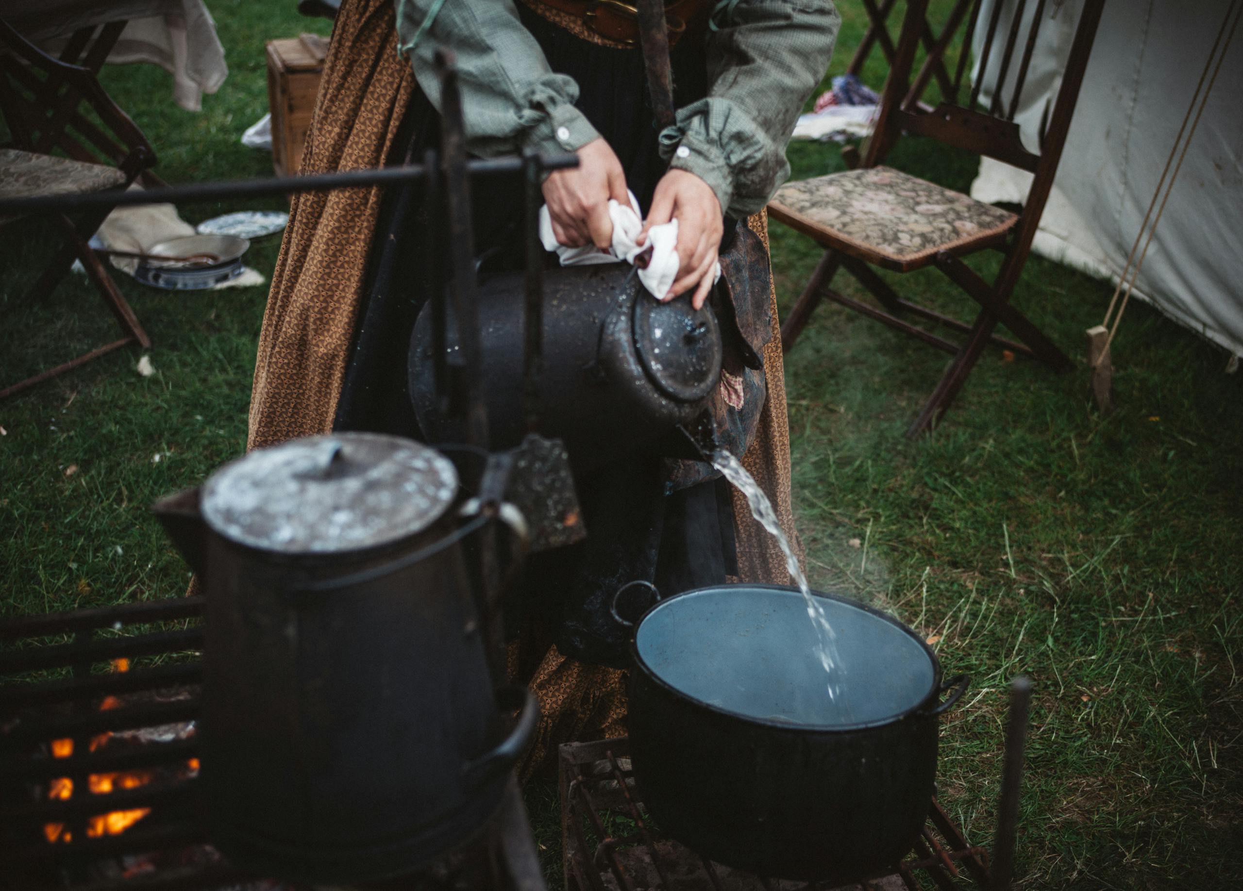Woman pouring water from a kettle into a pot outdoors during a camping setup. Warm, rustic scene.